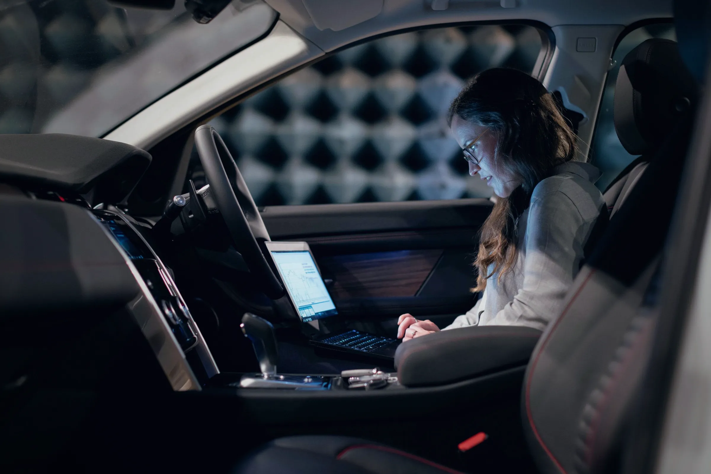 Test engineer in the driver's seat logging vehicle signals on a laptop inside a darkened acoustic test chamber