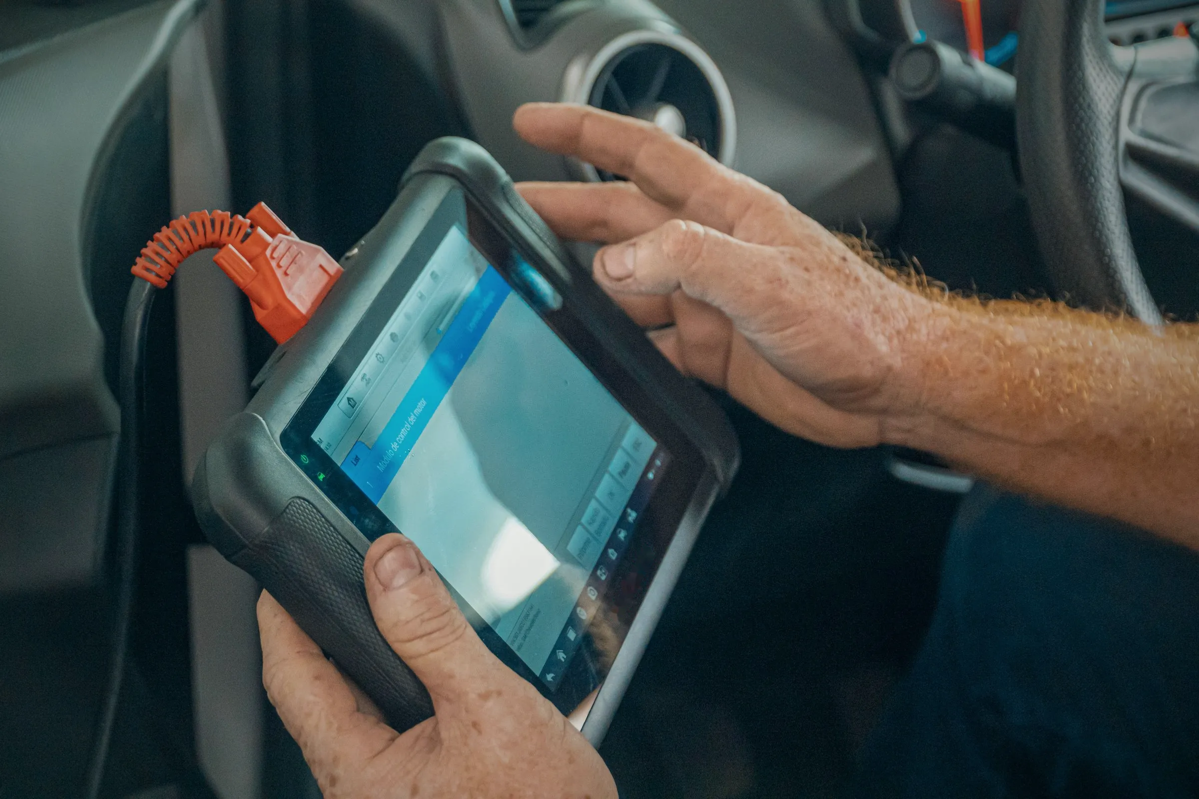 Technician's hands on a diagnostic tablet cabled into a car's diagnostic port